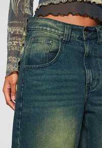 Džínové kalhoty v tmavě zelené barvě, s vysokým pasem, předními kapsami a bledým vzhledem. Šití podél švů.