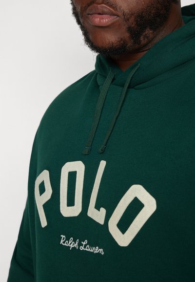 Zelená mikina s kapucí s nápisem "POLO" v smetanovém filcu a výšivkou "Ralph Lauren" pod ním. Tkanina vypadá měkce a má texturovaný povrch.