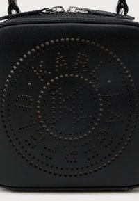 Svart teksturert veske med sirkulært perforert logodesign på fronten og en toppglidelås med lærhåndtak.