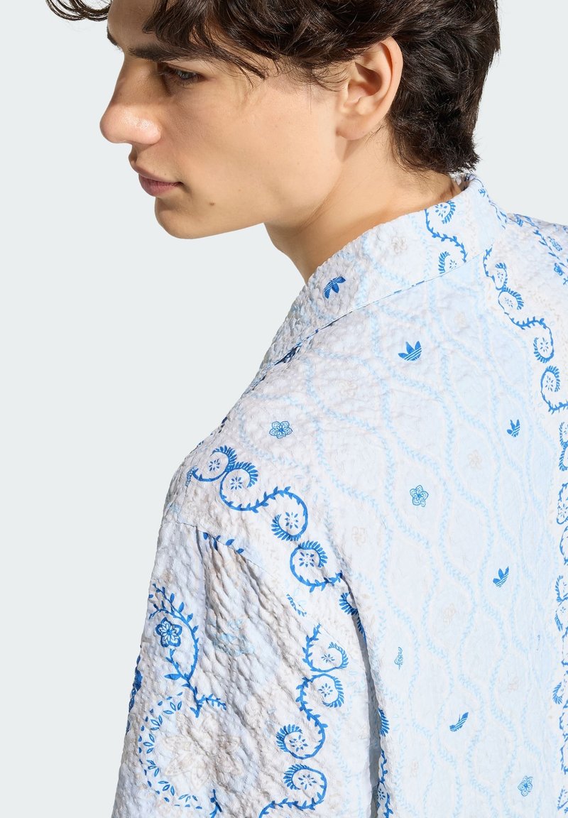 Jong persoon met krullend donker haar, gekleed in een wit gestructureerd shirt met blauwe bloemen- en spiraalpatronen, ziend naar links tegen een effen achtergrond.