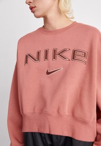 Sudadera Nike rosa con un corte oversized, puños y dobladillo acanalados. Presenta un gran logo bordado en colores contrastantes en la parte frontal.