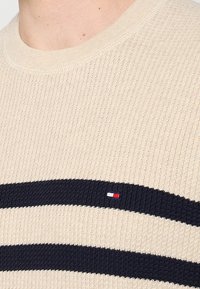 Maglione lavorato a maglia beige con strisce orizzontali blu navy, texture a coste e una piccola toppa con logo caratterizzata da dettagli rossi, bianchi e blu.
