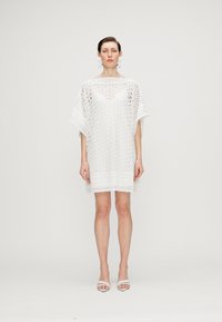 Rochie albă cu model din dantelă, cu mâneci largi și decolteu în V, având un material subtil texturat și o croială lejeră, purtată cu sandale albe.