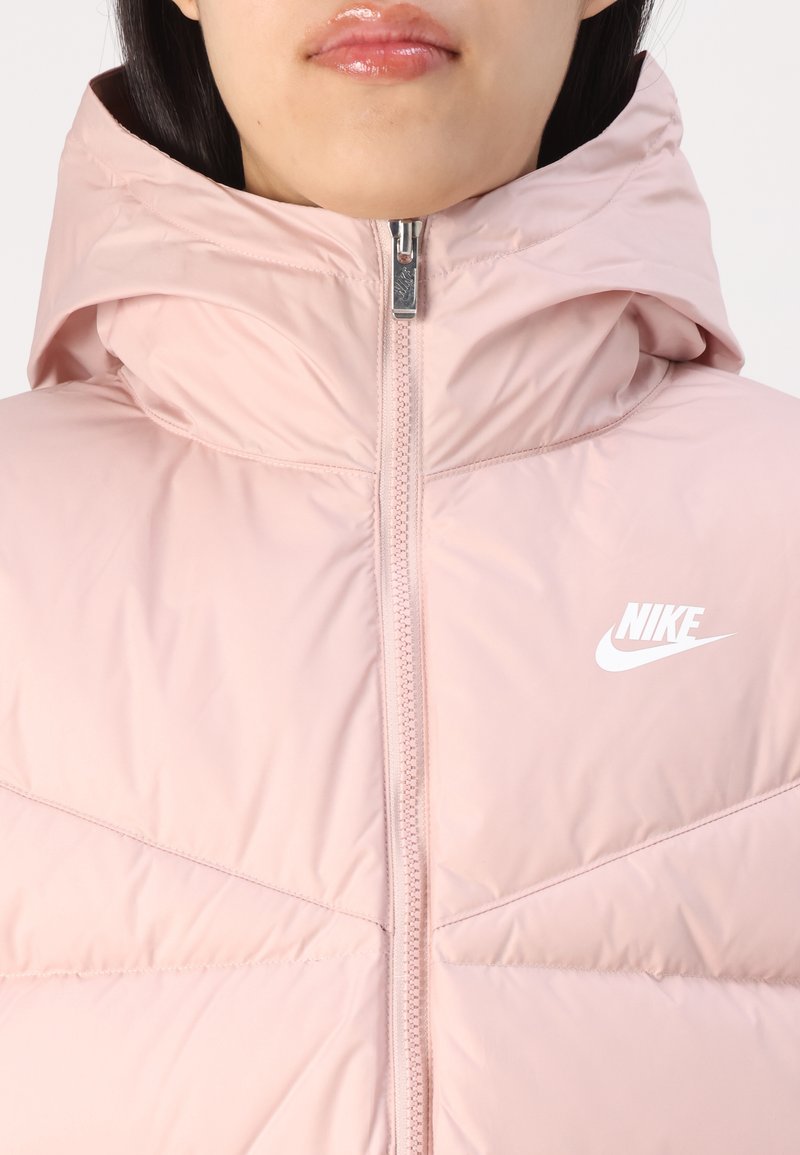 Rosa gepolsterte Jacke mit hohem Kragen und Kapuze. Vorderer Reißverschluss und weißes Nike-Logo auf der linken Brust. Glattes, leichtes Material.