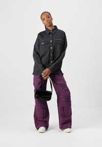 Camicia jacket nera oversized con tasche frontali e bottoni in ottone, abbinata a pantaloni cargo viola e una piccola borsa nera. Scarpe bianche.