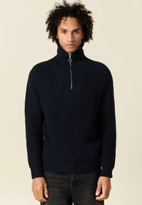 Pull marin côtelé avec col montant et fermeture éclair demi-longueur, arborant une surface texturée et des manches ajustées. Porté avec un jean foncé.