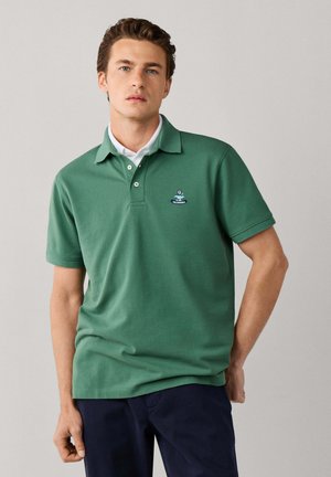 Joven con cabello castaño corto, vistiendo una camisa polo verde y pantalones oscuros, de pie frente a un fondo claro liso.
