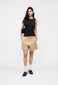 Μαύρο αμάνικο τοπ, μπεζ σορτς και μαύρα μοκασίνια με λευκές κάλτσες. Το μοντέλο κρατά μια μικρή μαύρη τσάντα χειρός. Μινιμαλιστικός σχεδιασμός σε λευκό φόντο.