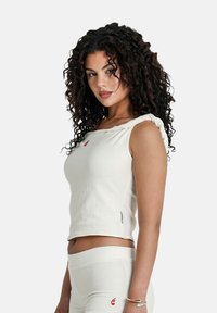 Witte cropped tanktop van een gestructureerde stof, met een gedraaide schouderband en een klein rood logo op de borst. Gecombineerd met bijpassende shorts.