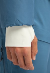 Niebieska rękawica kurtki z elastycznym mankietem, odsłaniająca białą wewnętrzną warstwę. Tekstura skóry na dłoni pokazuje naturalny kolor i detale.