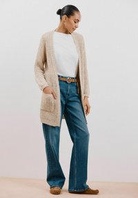 Bezešvý béžový cardigan s otevřeným předním dílem a kapsami, nošený přes bílou košili a modré džíny s vysokým pasem. Hnědý pásek a boty doplňují celé outfit.