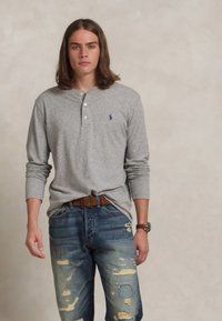 Νεαρός άνδρας με καστανά μαλλιά έως τους ώμους, φορώντας γκρι πουκάμισο με μακρύ μανίκι και φθαρμένα μπλε τζιν με καφέ ζώνη.