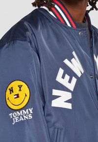 Granatowa kurtka bomberka z kołnierzem w paski w kolorach czerwonym, białym i niebieskim, białym napisem "NEW" oraz żółtą naszywką z uśmiechniętą buźką i tekstem "NY" oraz "TOMMY JEANS".