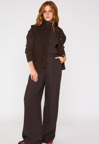 Gilet lavorato a maglia marrone con motivo a trecce, indossato sopra un dolcevita marrone scuro e pantaloni a palazzo grigi a vita alta. Accessori includono un bracciale.