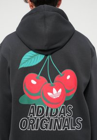 Pelēka kapuce ar lielu sarkano ķirsīšu grafiku un zaļu lapu. Apakšā atrodas teksts "ADIDAS ORIGINALS" treknā baltā fontā.