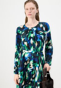 Vestuário de manga longa em azul, verde e preto com padrão abstrato, apresentando pregas e um cinto correspondente, combinado com uma bolsa de pele escura.