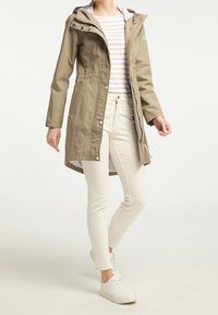Parka imperméable beige avec fermeture éclair frontale, boutons-pression et capuche. Comprend une doublure intérieure douce et des poches latérales. Portée sur un pantalon blanc.