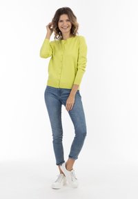 Cardigan botton-up verde lime con collo rotondo, polsini e orlo a coste, abbinato a jeans skinny blu e sneakers bianche.