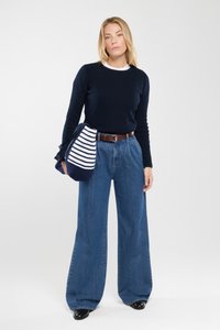 Marineblaues Sweatshirt, weißer Kragen, weite blaue Jeans, brauer Gürtel, schwarze Schuhe, hält eine marine- und weiß gestreifte Tasche aus strukturiertem Stoff.