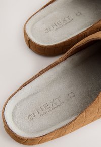 Chaussons en daim marron avec semelles intérieures rembourrées gris clair, présentant un marquage en relief « NEXT Forever Comfort » et des côtés texturés cousus.