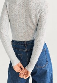 Lichtgrijze, gestructureerde gebreide trui met een chevronpatroon, gecombineerd met hoogwaisted blauwe denim jeans. Handen samengevoegd op de taille.