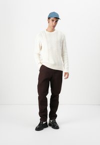 Creme striktrøje med strukturmønster, brune bukser, sorte sko og blå baseballkasket. Modellen står foran en hvid baggrund.