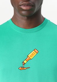 Grønn bomull t-skjorte med et grafisk motiv af en sauceskafter, der hælder orange sauce, med en gul label, der viser ordet "NIKE."
