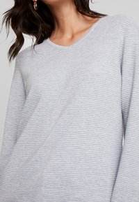 Vaaleanharmaa ribbineulottu pusero, jossa on V-aukko, pitkät hihat ja hienovaraiset tekstuurityksityiskohdat kankaassa.