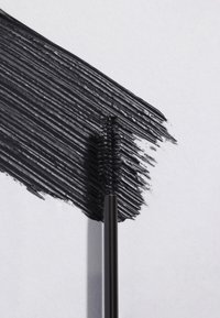 Czarna szczoteczka do tuszu do rzęs obok grubej, czarnej kreski tuszu na białym papierze, z teksturowanym i błyszczącym wykończeniem.