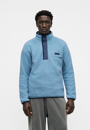 Homme face à l’objectif, portant un pull polaire bleu clair avec col boutonné et poche poitrine zippée, sur un pantalon gris, fond clair uni.