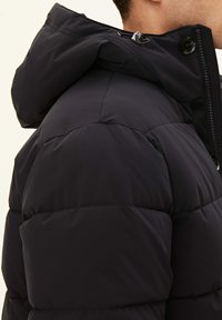 Svart puffjacka med huva, horisontell quiltad design, dragkedjestängning och tryckknappdetaljer. Slät textur, isolerad för värme.