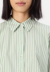Grün-weiß gestreifte Bluse mit klassischem Kragen, ausgestattet mit Knopfverschlüssen und leichtem Stoff. Nahaufnahme zeigt Textur und Muster.