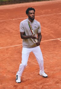 Tenisový hráč v světle šedém polu s límečkem, bílých sportovních kalhotách a barevným tenisovým raketou, na antukovém kurtu.