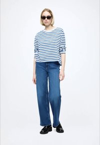 Striped blue and white long-sleeve shirt, blue wide-leg jeans, and black platform shoes. Model wears dark sunglasses. Simple background.