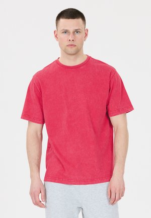 Ung mand med kort hår iført en ensfarvet, falmet rød T-shirt og lysegrå bukser, der står mod en hvid baggrund.