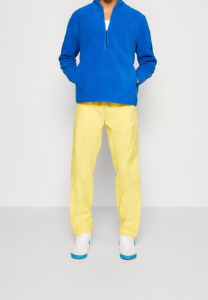 Person wearing a bright blue quarter-zip fleece, pale yellow pants, and white sneakers with blue soles, standing against a plain white background.