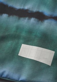 Textilprodukt med en abstrakt blågrön och svart gradientdesign, med en texturerad yta och en rektangulär silveretikett med rutmönster.