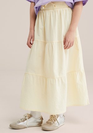 Ženska, oblečena v bledo rumeno večnivojsko krilo, srebrne superge, bele nogavice in vijoličasto majico, stoji pred nevtralnim ozadjem.