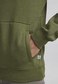 Sweatshirt vert avec une finition texturée, doté d'une poche kangourou, de poignets côtelés et d'une étiquette sur le côté. Tissu doux en mélange de coton.
