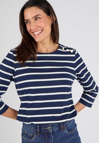 Maglietta a righe blu navy e bianca con scollo rotondo, maniche a tre quarti e tre bottoni decorativi sulla spalla sinistra. Realizzata in tessuto morbido.