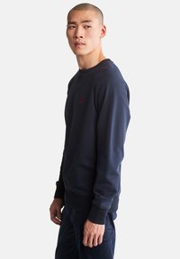 Felpa girocollo blu navy con polsini e orlo a costine, caratterizzata da un piccolo logo rosso sul petto e un tessuto liscio e testurizzato.