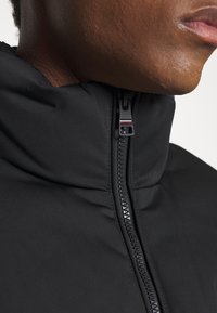 Gros plan sur le cou et le menton d'une personne portant une veste noire zippée jusqu'au col, sur un fond uni.