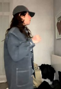 Άτομο με γκρι μπουφάν και καπέλο στέκεται μέσα κοντά σε έναν τοίχο με ένα κορνιζαρισμένο πόστερ και σωρούς ρούχων στο πάτωμα.
