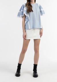 Una donna è in piedi con una camicia blu con pizzi, una mini gonna bianca e stivali da combattimento neri su uno sfondo bianco.