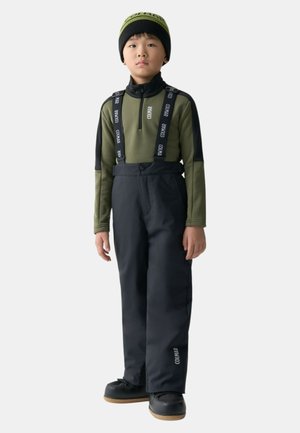 Pantaloni impermeabili neri con bretelle regolabili e top verde a maniche lunghe. Presentano dettagli con logo e un design aderente. Stivali neri.