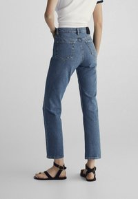 Jeans in denim blu con vestibilità dritta, vita alta e cuciture a contrasto; presentano due tasche posteriori e una texture liscia. Indossati con sandali neri.