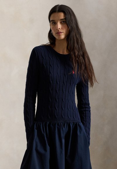 Μπλε σκούρο πουλόβερ με πλέξη καλώδιο και μακριά μανίκια, με λεπτομέρεια σιέντας με πιέτες στο τελείωμα. Μικρό κόκκινο λογότυπο στο στήθος.
