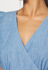 Jasnoniebieska tkanina chambray z głębokim dekoltem w kształcie litery V, z krótkimi rękawami i miękką, teksturowaną powierzchnią. Brak widocznych wzorów lub elementów metalowych.