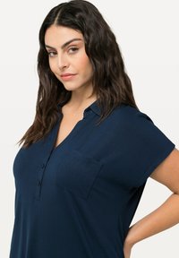 Mörkblå kortärmad skjorta med krage, knappslå och en enda bröstficka. Slät textur, avslappnad passform.