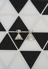 Silver triangulära örhängen med tre inlagda klara stenar längs en diagonal på en texturerad bakgrund. Mot en geometrisk tile-mönster.